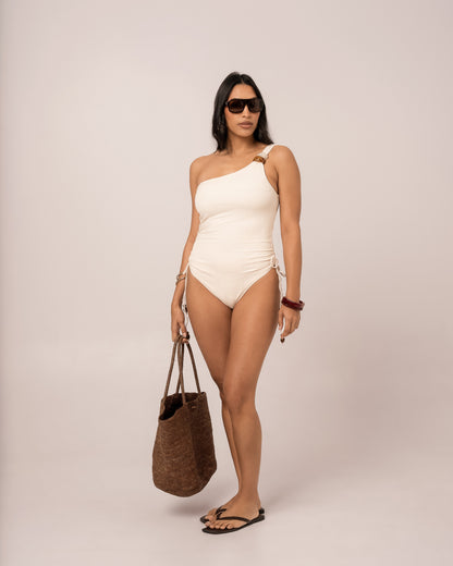 Ivory one shoulder one piece swimsuit styled with a woven raffia tote, showing the adjustable ruched side tie detail — honeymoon resortwear
