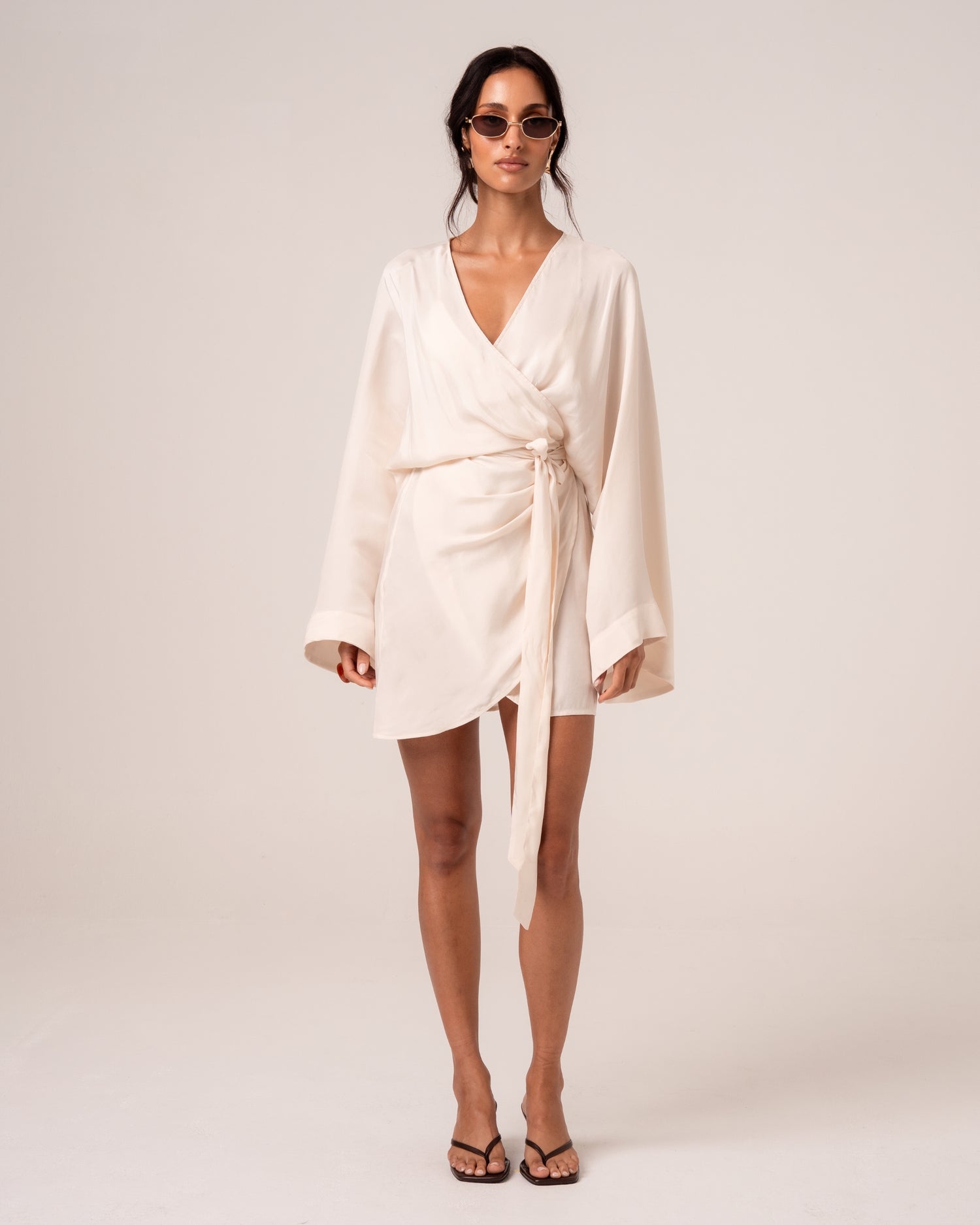 Model wearing an ivory batwing wrap mini dress with a deep V-neckline, wide sleeves and side tie, studio front view