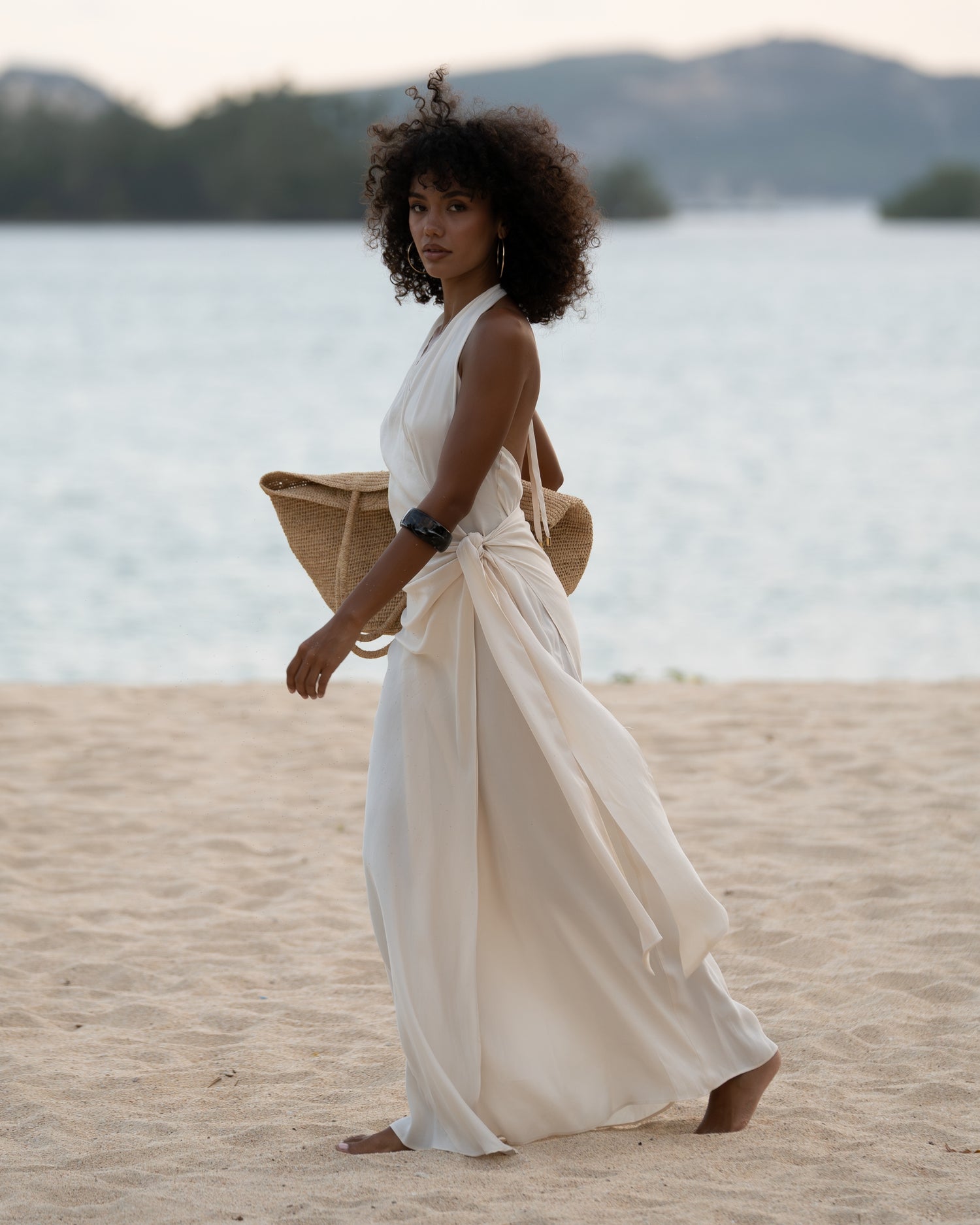 Woman walking on the beach in an ivory halter wrap maxi dress with cascading side tie, carrying a straw tote