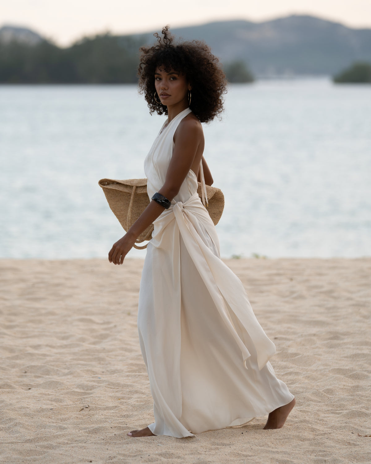 Woman walking on the beach in an ivory halter wrap maxi dress with cascading side tie, carrying a straw tote