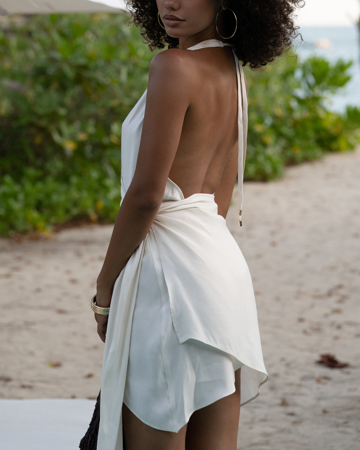 Close back detail of an ivory halter wrap mini dress with open back and gold-tipped tie ends, worn at the beach