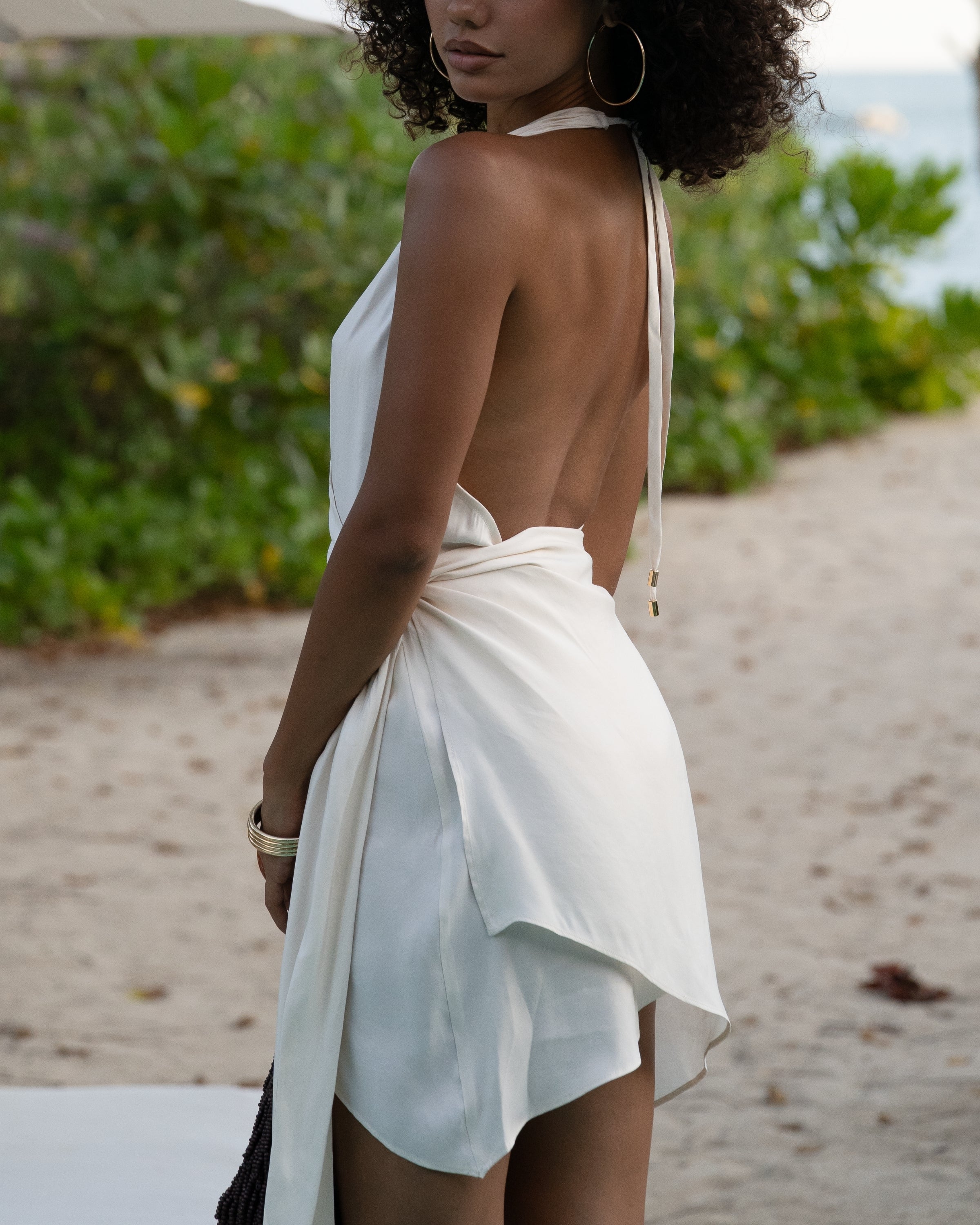 Close back detail of an ivory halter wrap mini dress with open back and gold-tipped tie ends, worn at the beach