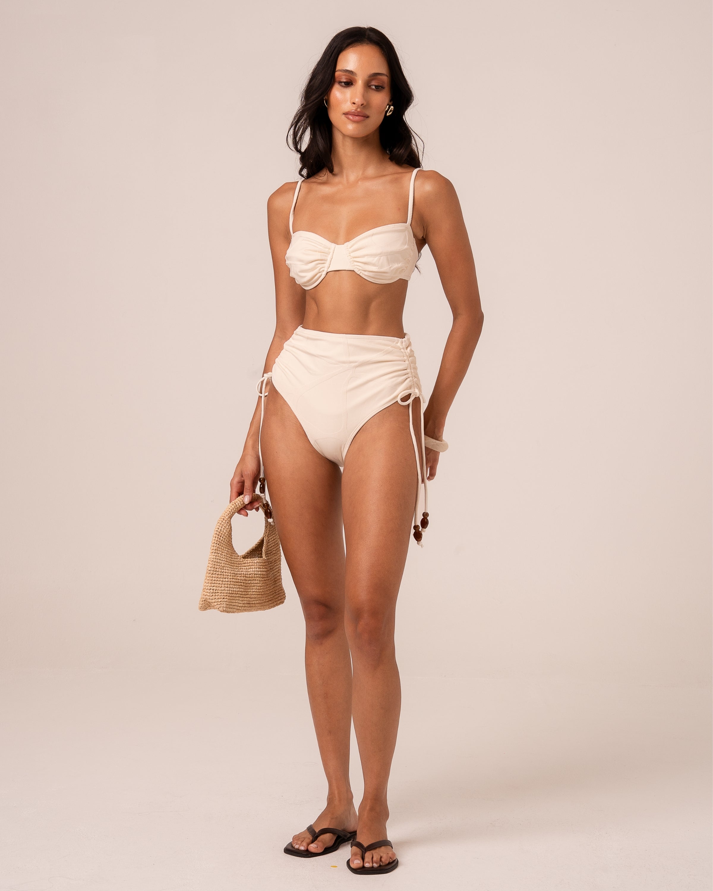 Full-length front view of an ivory high waist tie side bikini bottom with beaded adjustable ties, styled with a bandeau top
