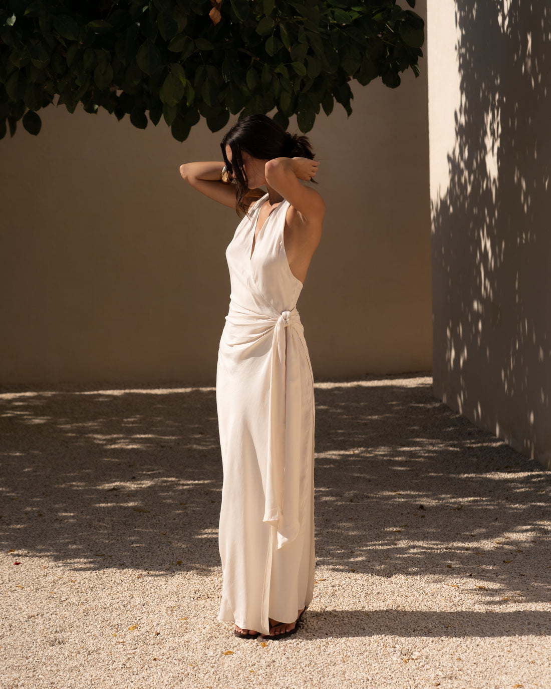 Side view of an ivory halter maxi dress with a cascading waist tie drape in a sunlit courtyard