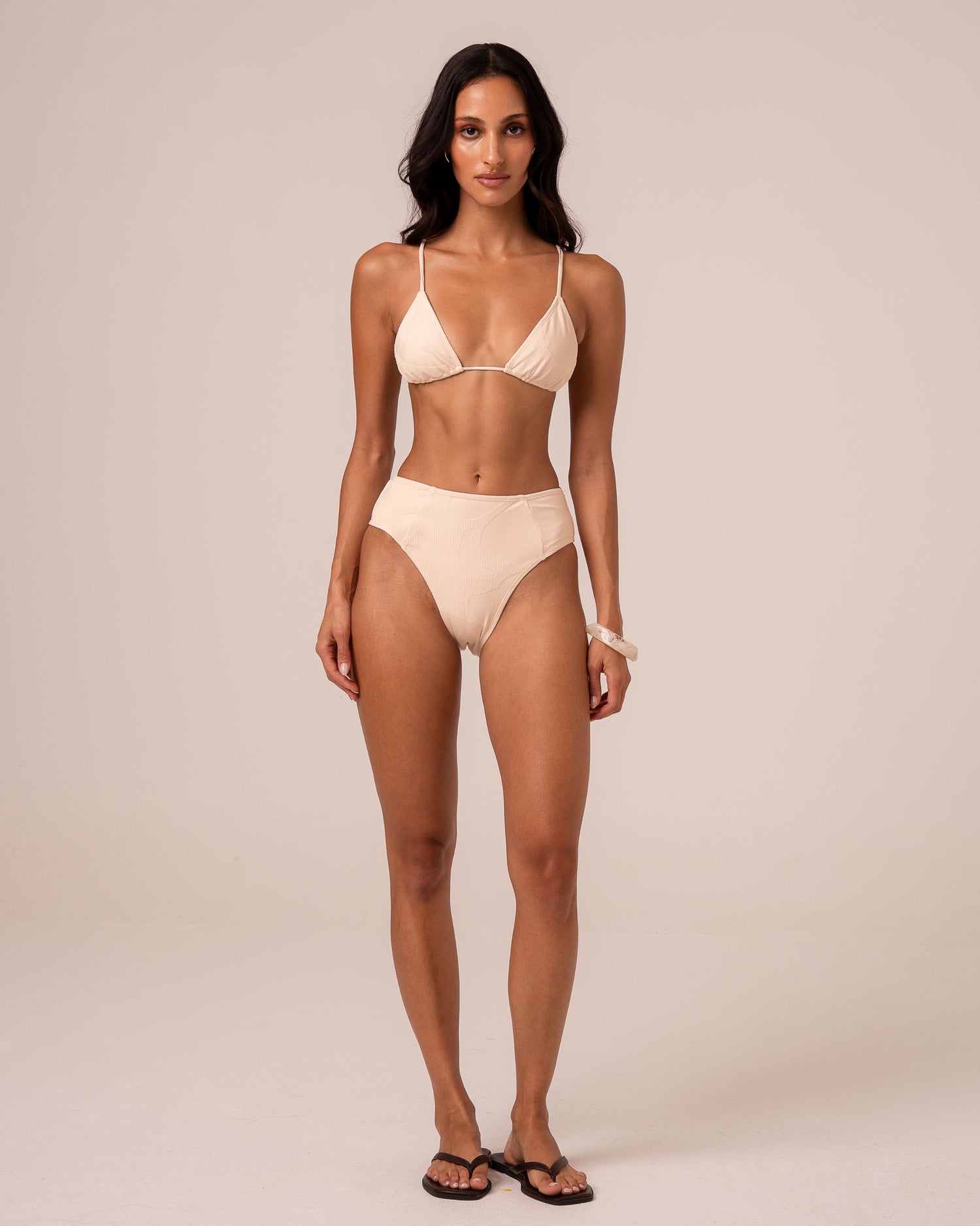 Model wearing an ivory mid rise bikini bottom with spliced panel seams and a higher leg, styled with a triangle top — studio front view