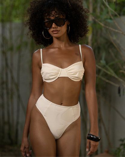 Ivory mid rise bikini bottom with spliced seams and a higher-cut leg, front view at a tropical resort