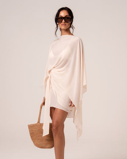Ivory resort cape knotted at the hip over a white mini skirt, styled with a straw tote — bridal resortwear styling