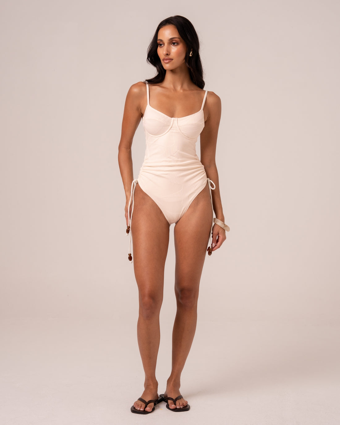 Model wearing an ivory underwire one piece swimsuit with thin adjustable straps and beaded ruched side ties, studio front view