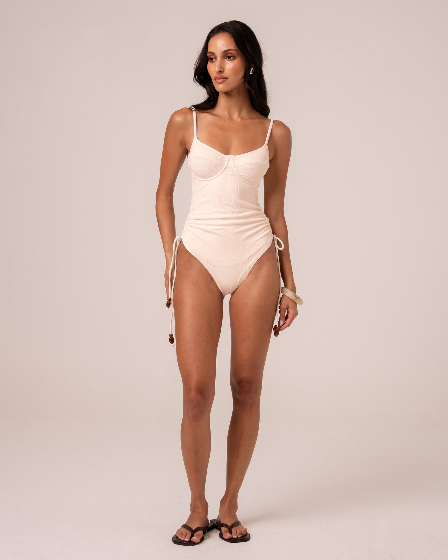 Model wearing an ivory underwire one piece swimsuit with thin adjustable straps and beaded ruched side ties, studio front view