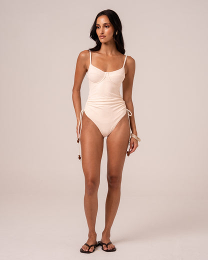 Model wearing an ivory underwire one piece swimsuit with thin adjustable straps and beaded ruched side ties, studio front view