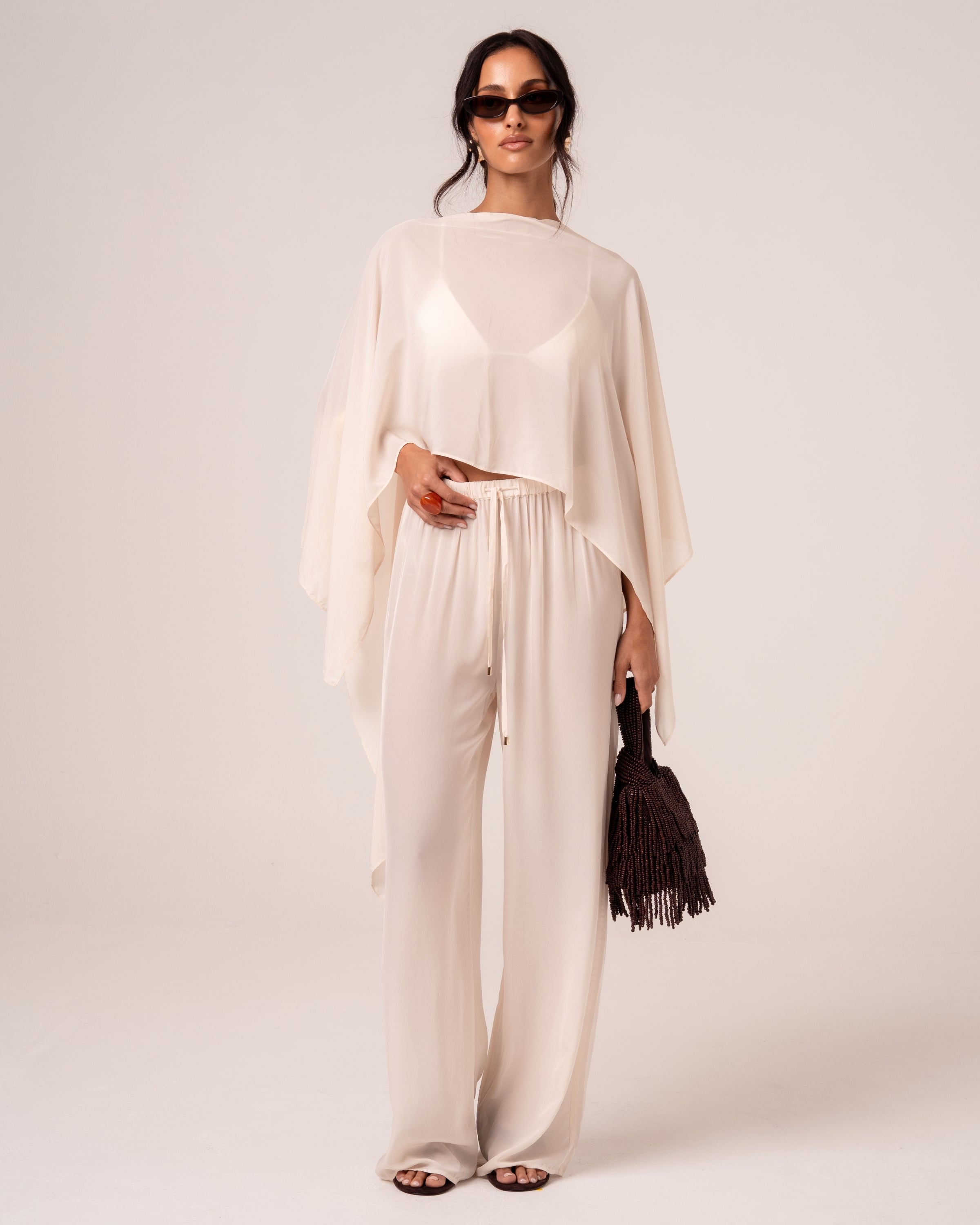 Model wearing ivory wide-leg resort pants with an elasticated drawstring waist and a matching sheer cape, front view