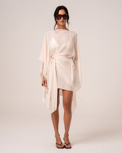 Front view of an ivory viscose wrap mini skirt with hip tie, styled with a sheer cape over a matching bikini