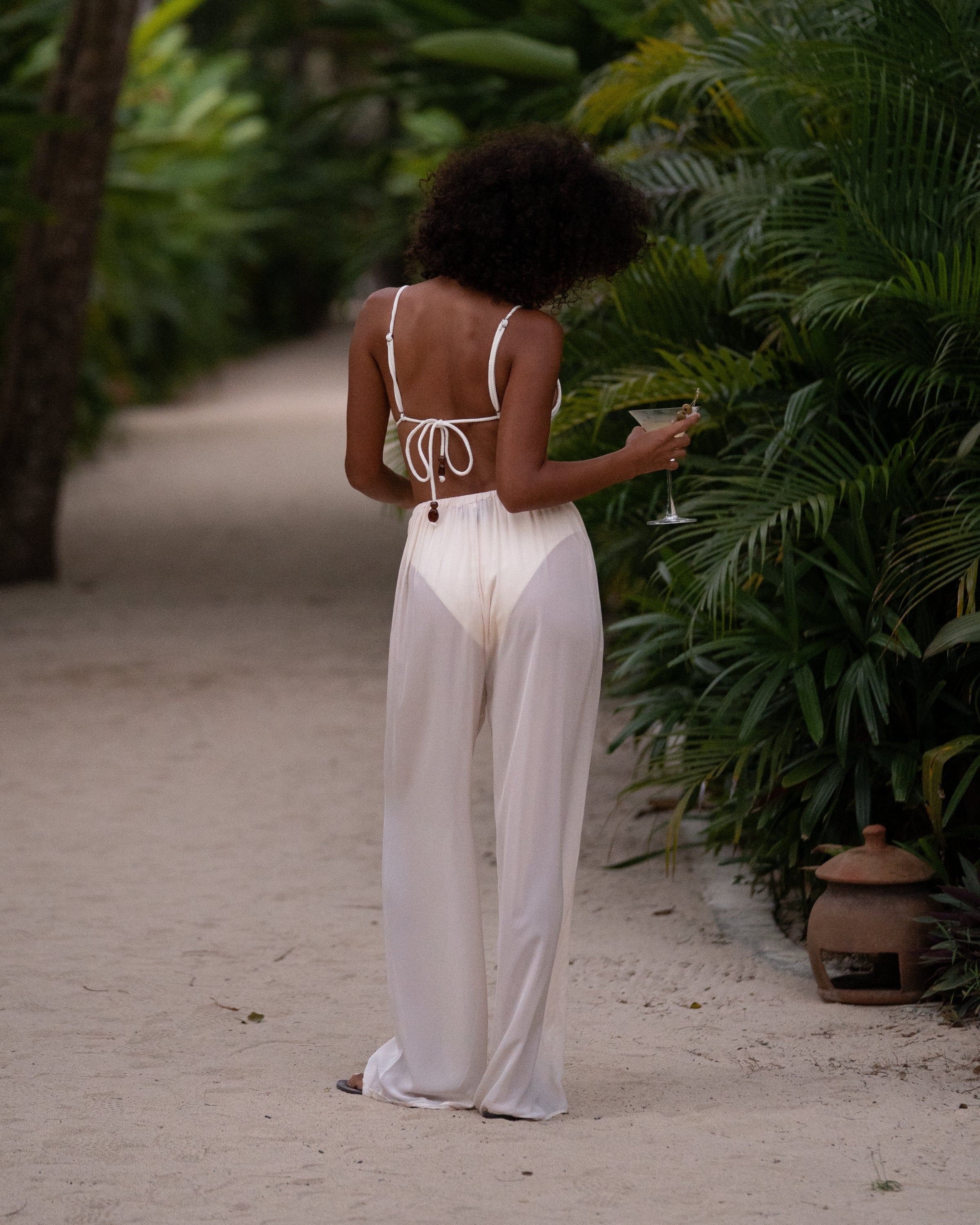 Back view of sheer ivory wide-leg resort pants worn over a white bikini on a palm-lined resort path