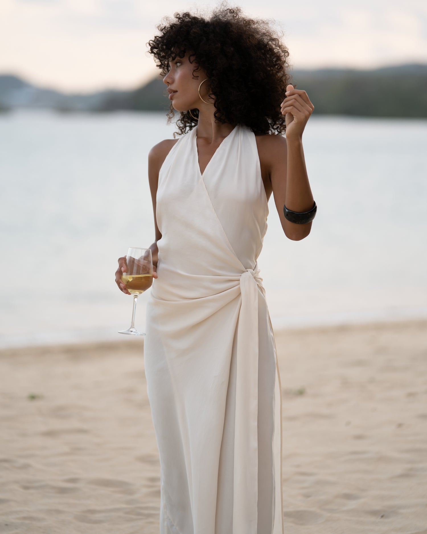 Close view of an ivory wrap maxi dress showing the plunging halter neckline and knotted side waist tie, ocean behind
