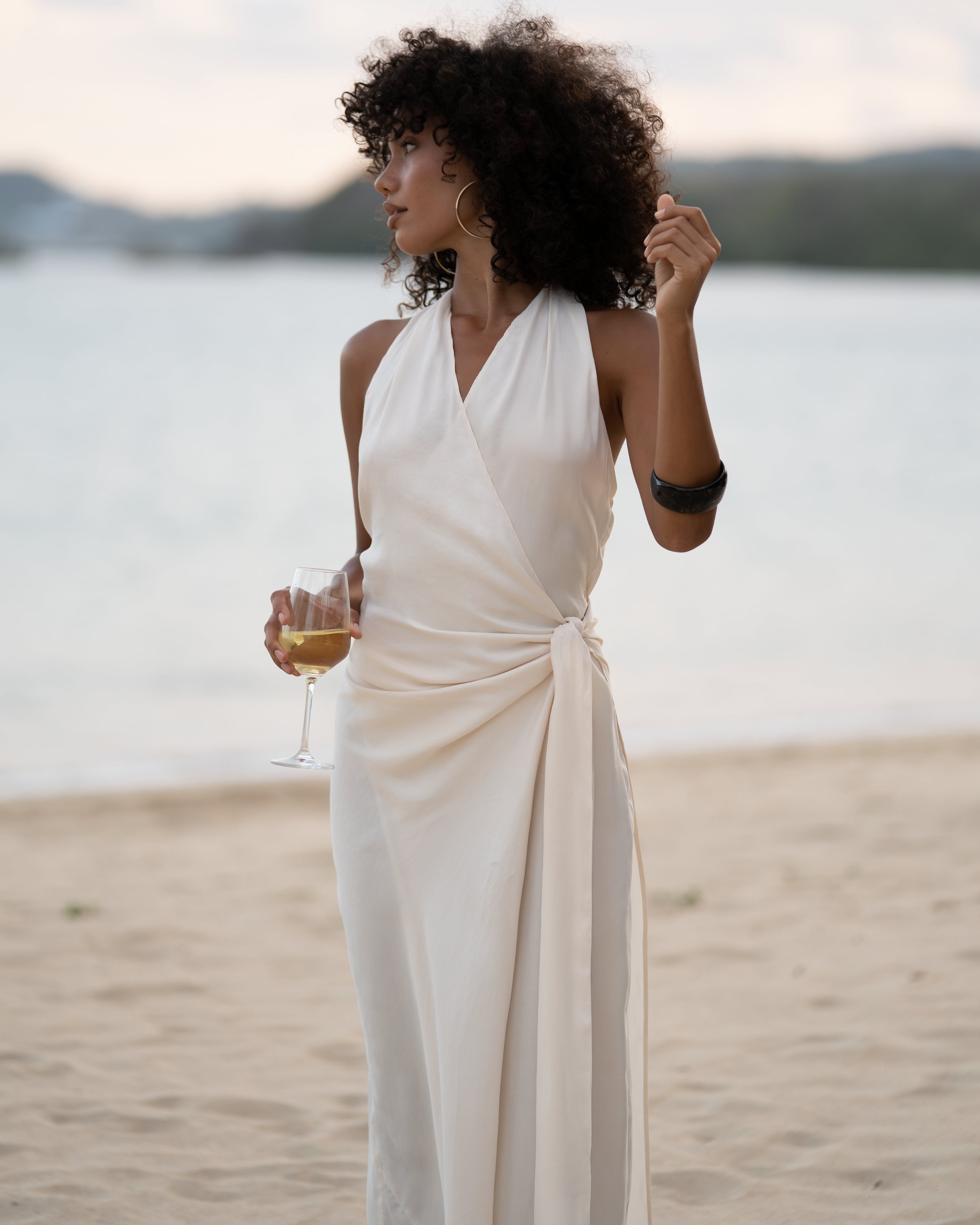 Close view of an ivory wrap maxi dress showing the plunging halter neckline and knotted side waist tie, ocean behind