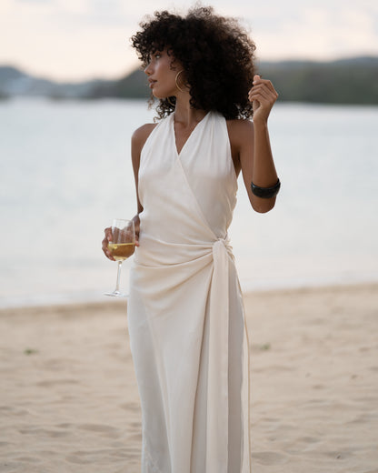 Close view of an ivory wrap maxi dress showing the plunging halter neckline and knotted side waist tie, ocean behind
