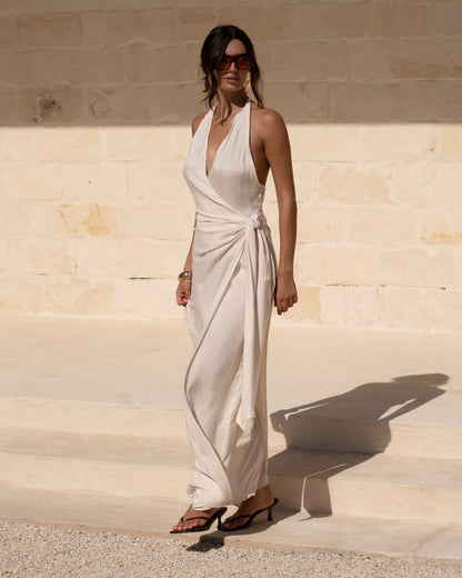 Ivory halter wrap maxi dress with plunging V-neckline and side knot worn with heels against a limestone wall — wedding guest resortwear
