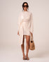 Model wearing an ivory wrap mini skirt tied at the hip, layered under a matching sheer resort cape with a straw bucket bag