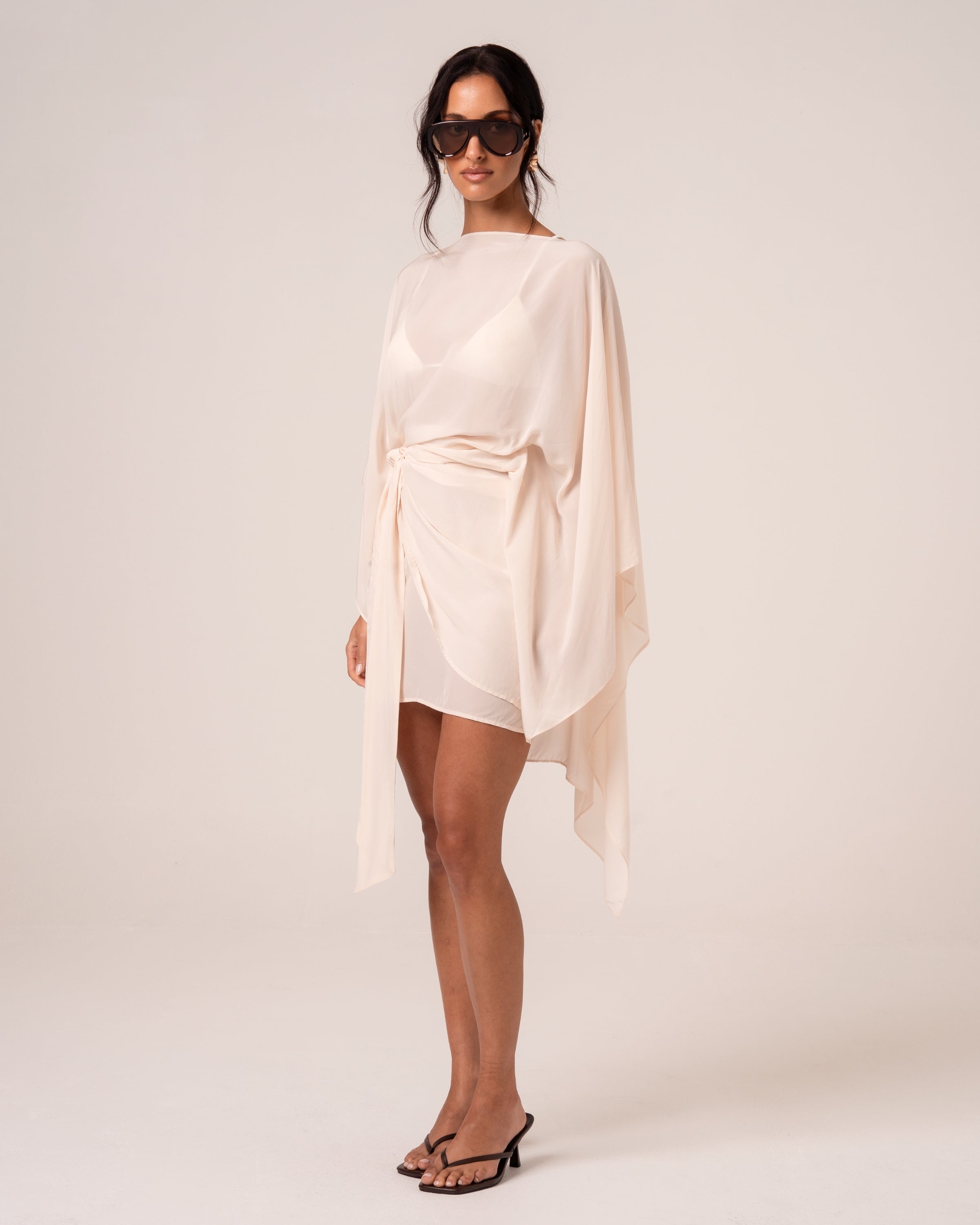 Side view of an ivory wrap mini skirt with a draped self-tie, worn with a sheer resort cape — elevated resortwear