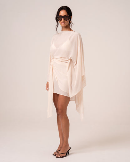 Side view of an ivory wrap mini skirt with a draped self-tie, worn with a sheer resort cape — elevated resortwear