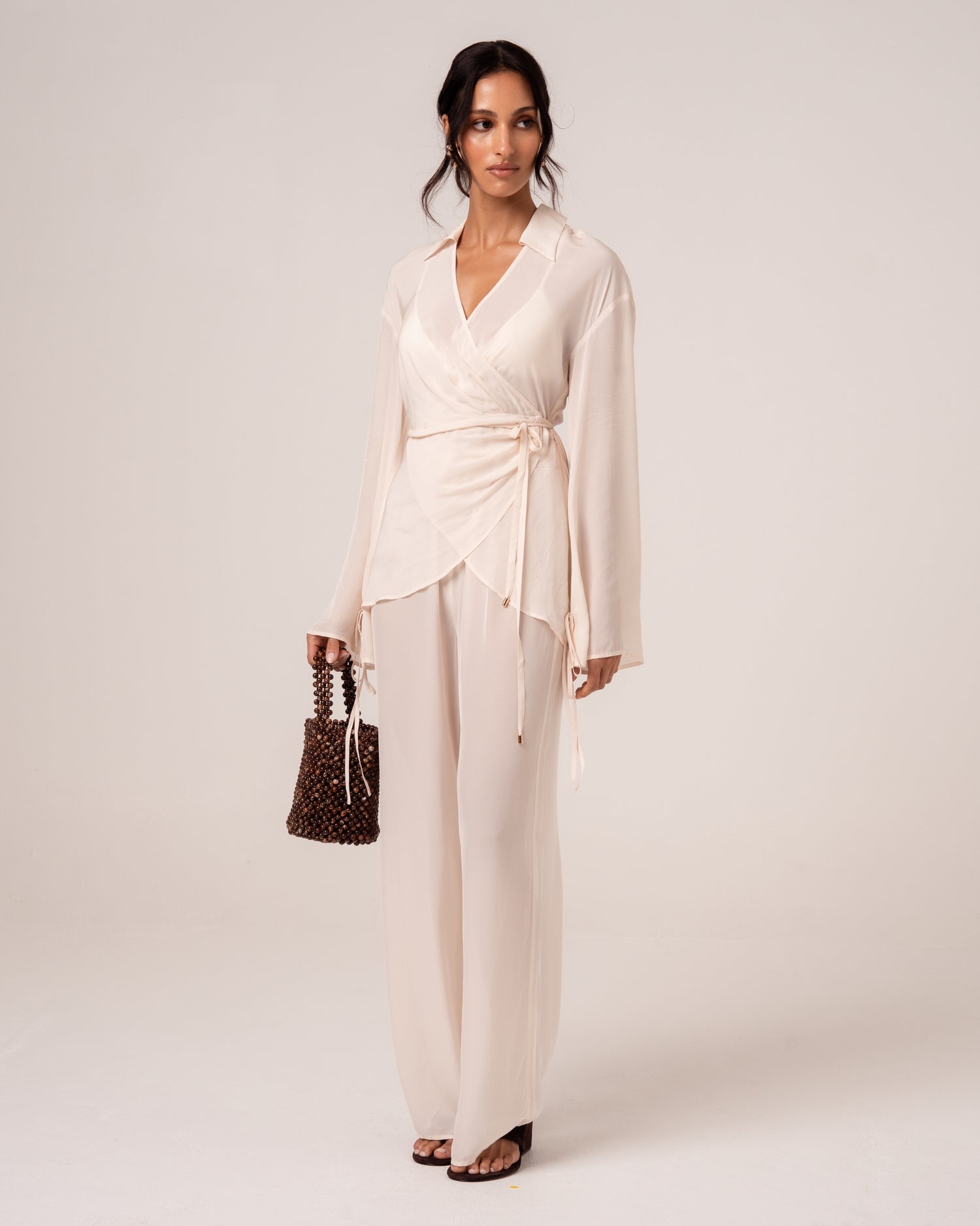 Side view of an ivory wrap shirt with waist tie worn with flowy wide-leg pants and a beaded bag