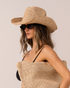 Model wearing a natural raffia straw cowboy hat with a wide curved brim, front angle, paired with a matching woven straw tote