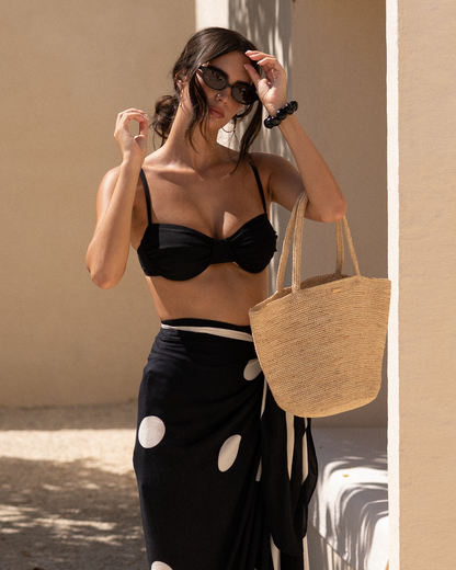 Natural raffia straw mini tote hanging from the forearm, close-up of model in a polka-dot sarong adjusting her sunglasses