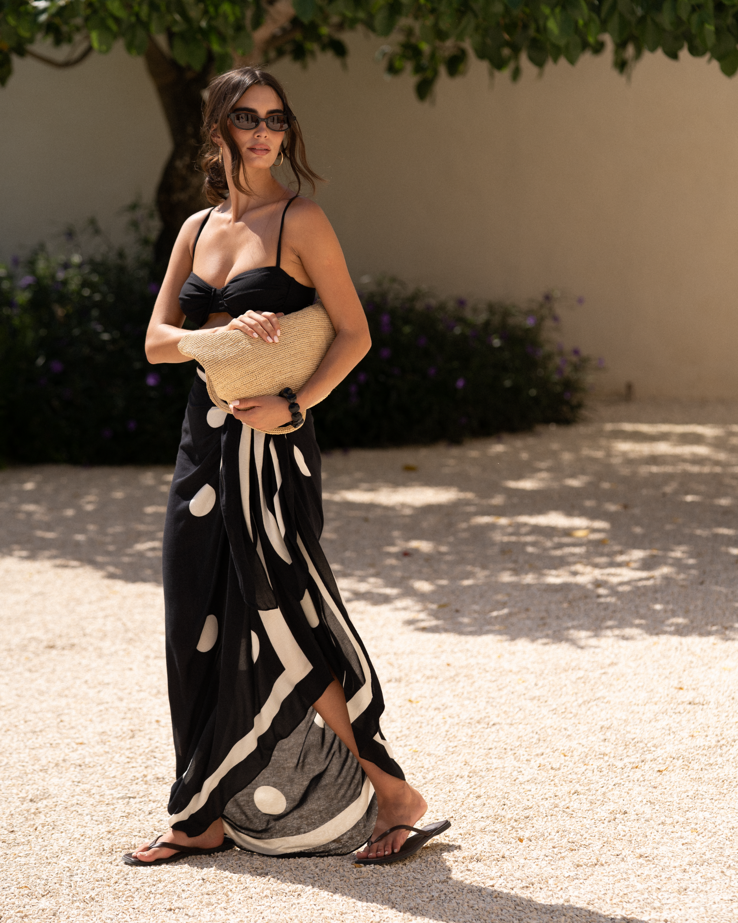 Natural raffia straw mini tote held against the torso with both hands, model in a polka-dot sarong standing beneath a leafy tree