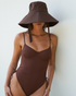 Model wearing the Ochre Lane Sun Hat in chocolate cotton, featuring a wide brim and tie straps, styled with the matching chocolate rib one-piece swimsuit.