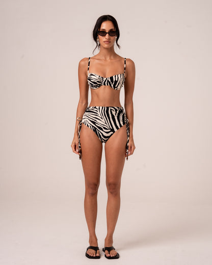 Model wearing zebra print high-waisted bikini bottoms with ruched side ties and wooden bead accents, paired with a matching bralette top, studio front view