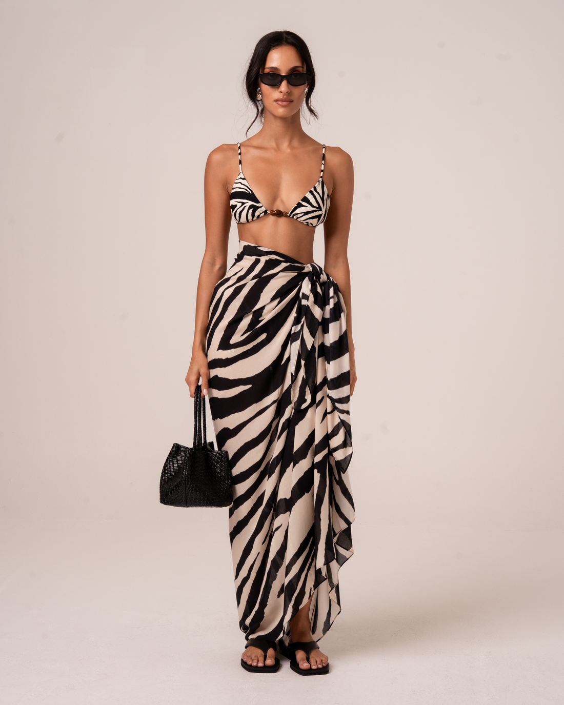 Model wearing a zebra print sarong wrap skirt with a front knot tie, styled over a matching triangle bikini top and a woven tote bag, studio front view