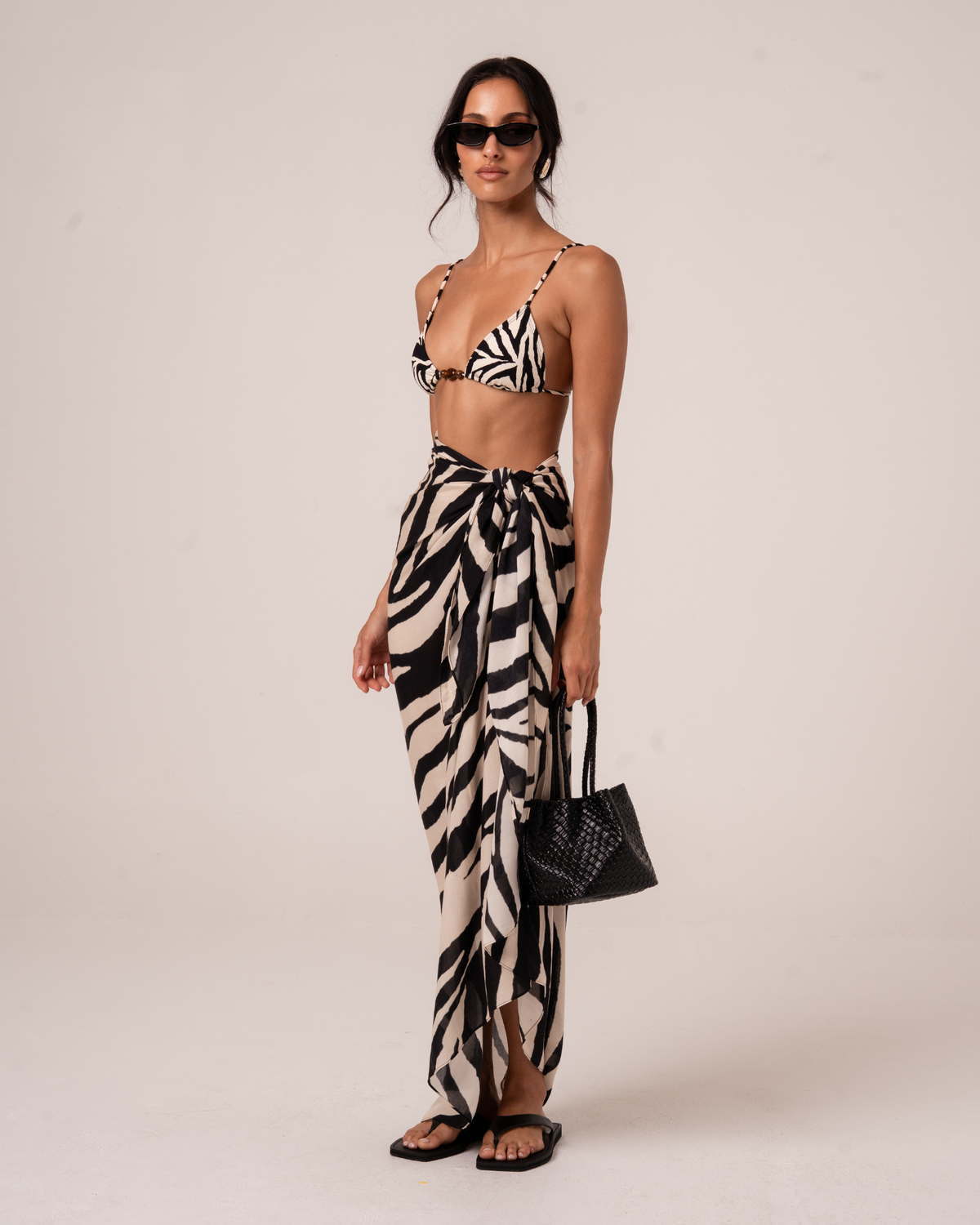 Three-quarter view of a zebra print sarong wrap skirt knotted at the waist, styled with a woven tote bag and matching bikini top