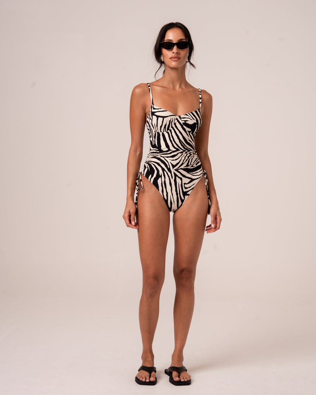 Model wearing a zebra print one-piece swimsuit with a sweetheart underwire neckline, thin adjustable straps, and a ruched side tie waist, studio front view