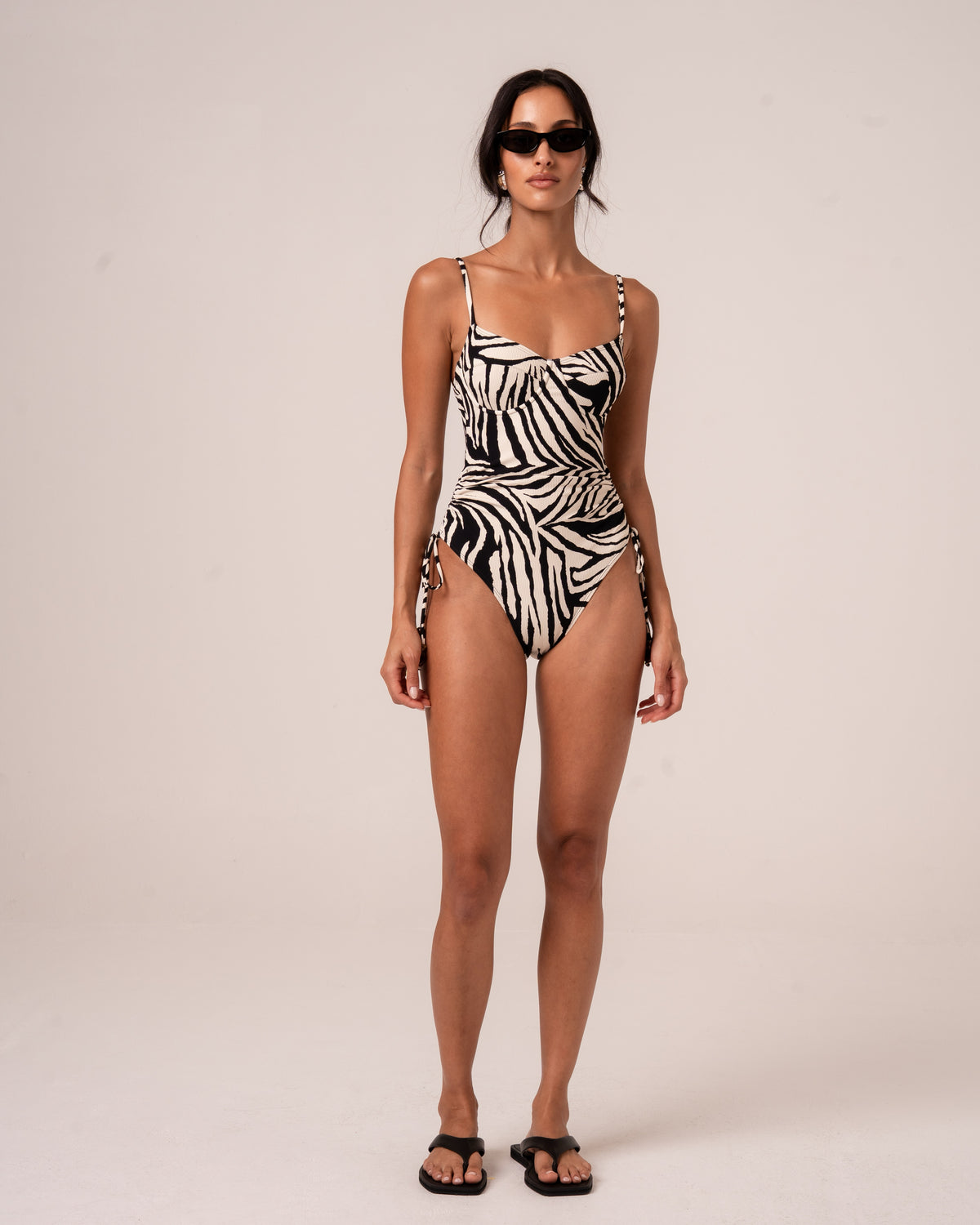 Model wearing a zebra print one-piece swimsuit with a sweetheart underwire neckline, thin adjustable straps, and a ruched side tie waist, studio front view