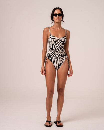Model wearing a zebra print one-piece swimsuit with a sweetheart underwire neckline, thin adjustable straps, and a ruched side tie waist, studio front view